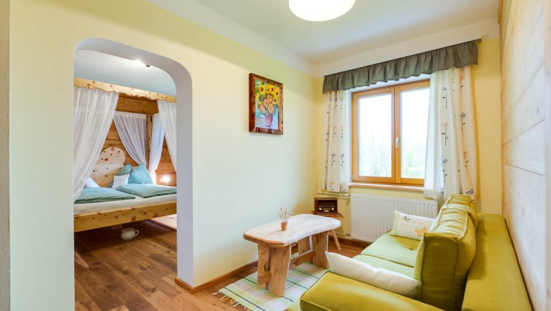 Rooms at the Höchbauernhof, © zVg Cozy room with wooden furniture, green sofa, large window and a four-poster bed in the background.