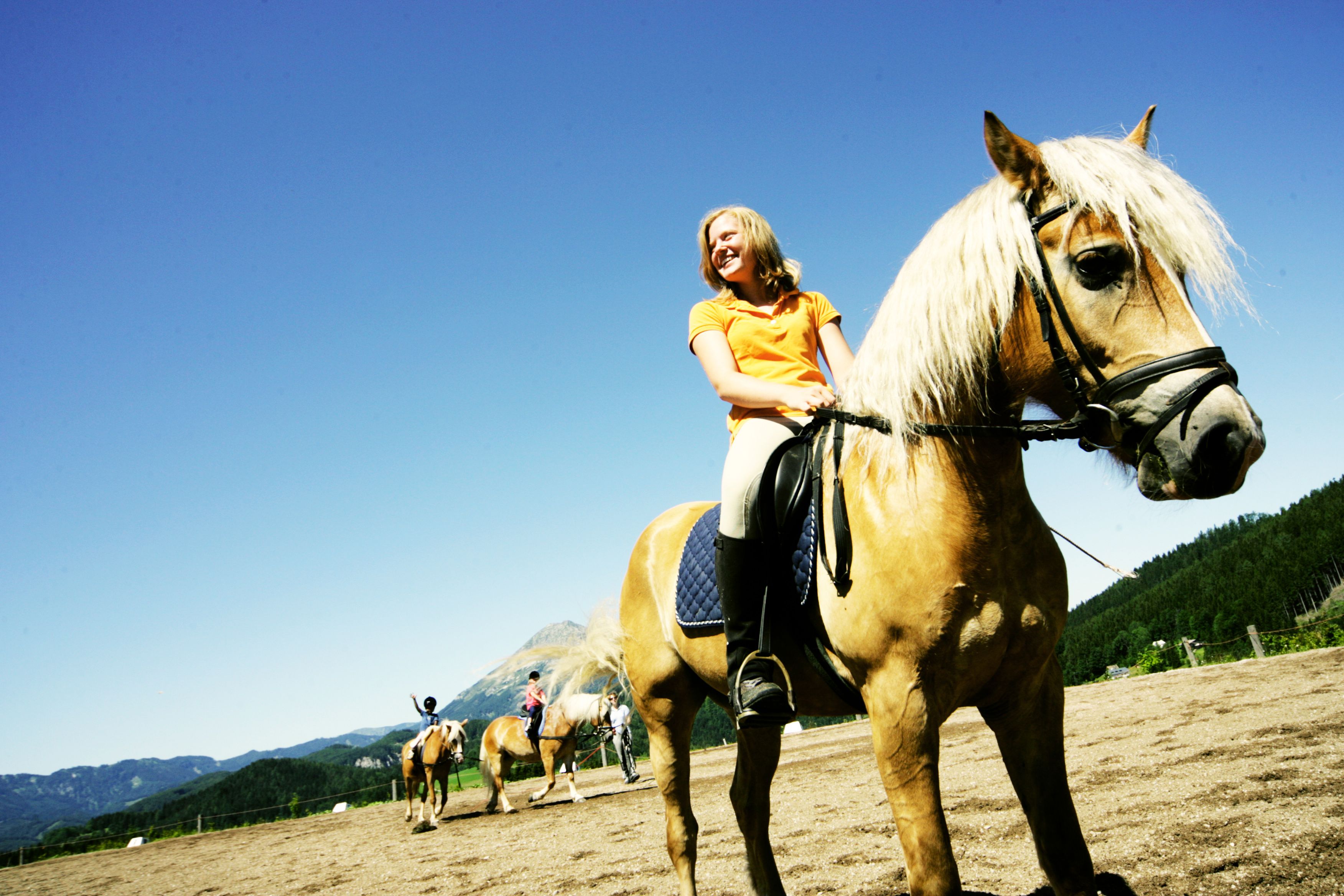 A woman rides a brown horse with a blond mane in a riding arena surrounded by mountains and blue sky.