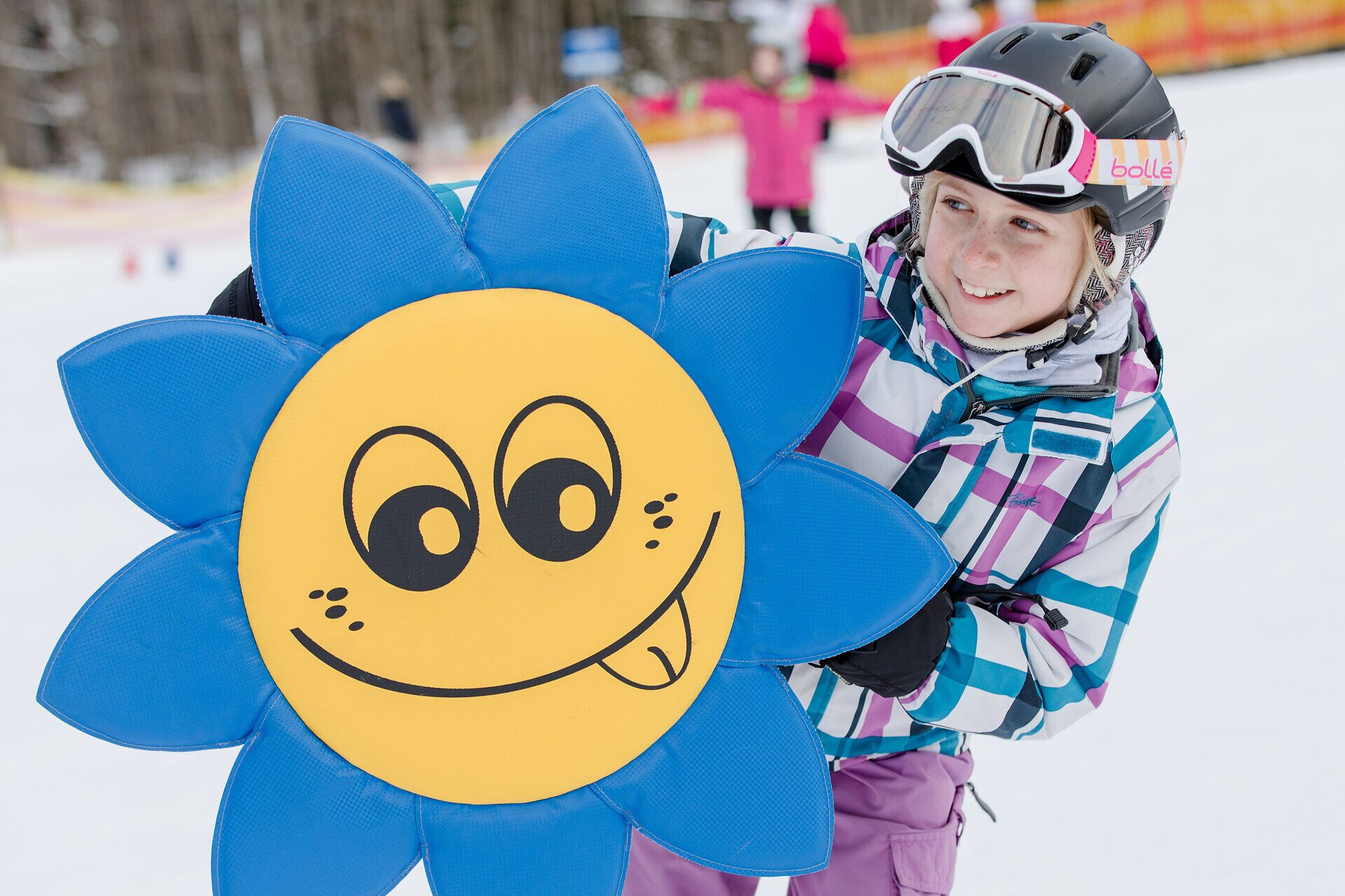 Ein fröhliches Kind strahlt beim Spielen im Schnee und hält ein buntes Sonnensymbol in den Händen. Die winterliche Landschaft um ihn herum ist mit schneebedeckten Bäumen geschmückt, während andere Kinder im Hintergrund fröhlich herumtollen. Hier wird der Winter zum unvergesslichen Erlebnis für die ganze Familie.