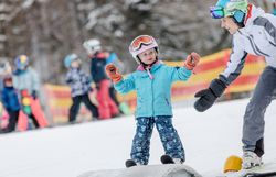Ein fröhliches Kind steht auf den Skiern, während ein geduldiger Lehrer ihm zur Seite steht. Umgeben von schneebedeckten Bergen und anderen Skifahrern, strahlt die Szene eine warme, einladende Atmosphäre aus, die die Freude am Wintersport für die ganze Familie verkörpert.
