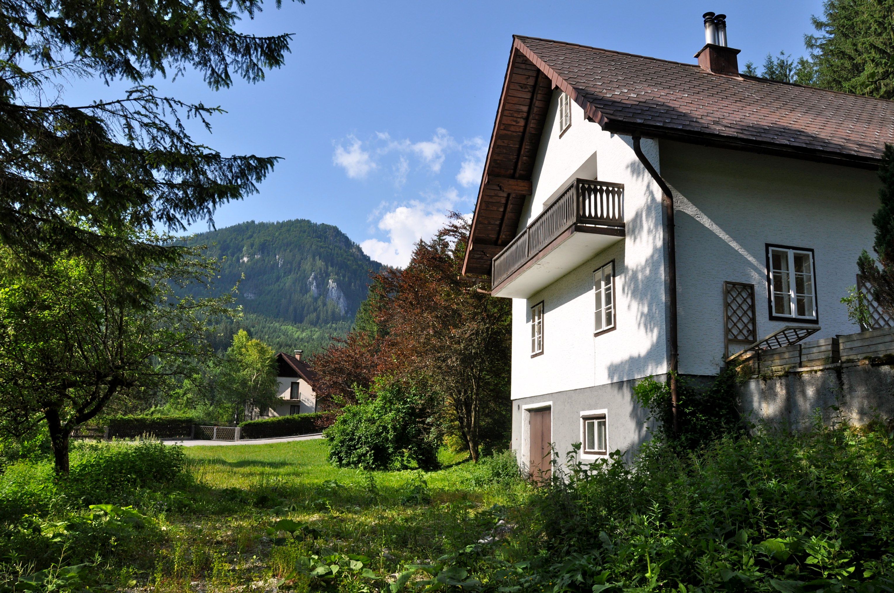 A white vacation home with balcony in front of a mountain landscape and green garden.