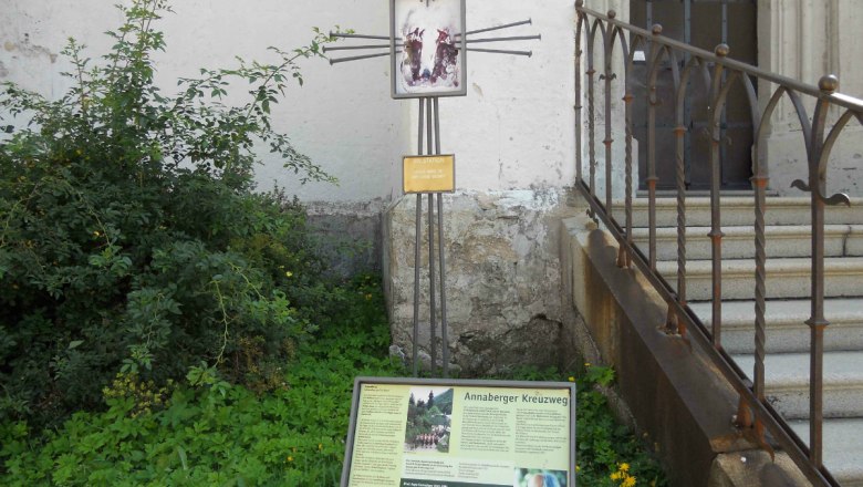 A sign with information about the Annaberg Way of the Cross next to a staircase and plants.