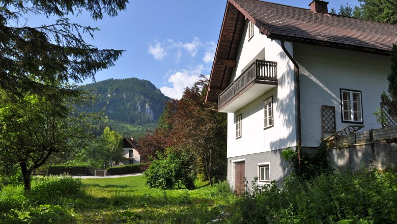 Ein wei&szlig;es Ferienhaus mit Balkon vor einer Berglandschaft und gr&uuml;nem Garten.