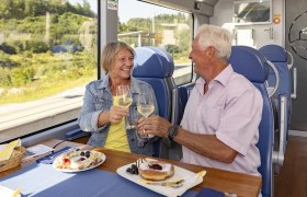 Ein älteres Paar stößt mit Weingläsern in einem Zug an, mit Essen auf dem Tisch.