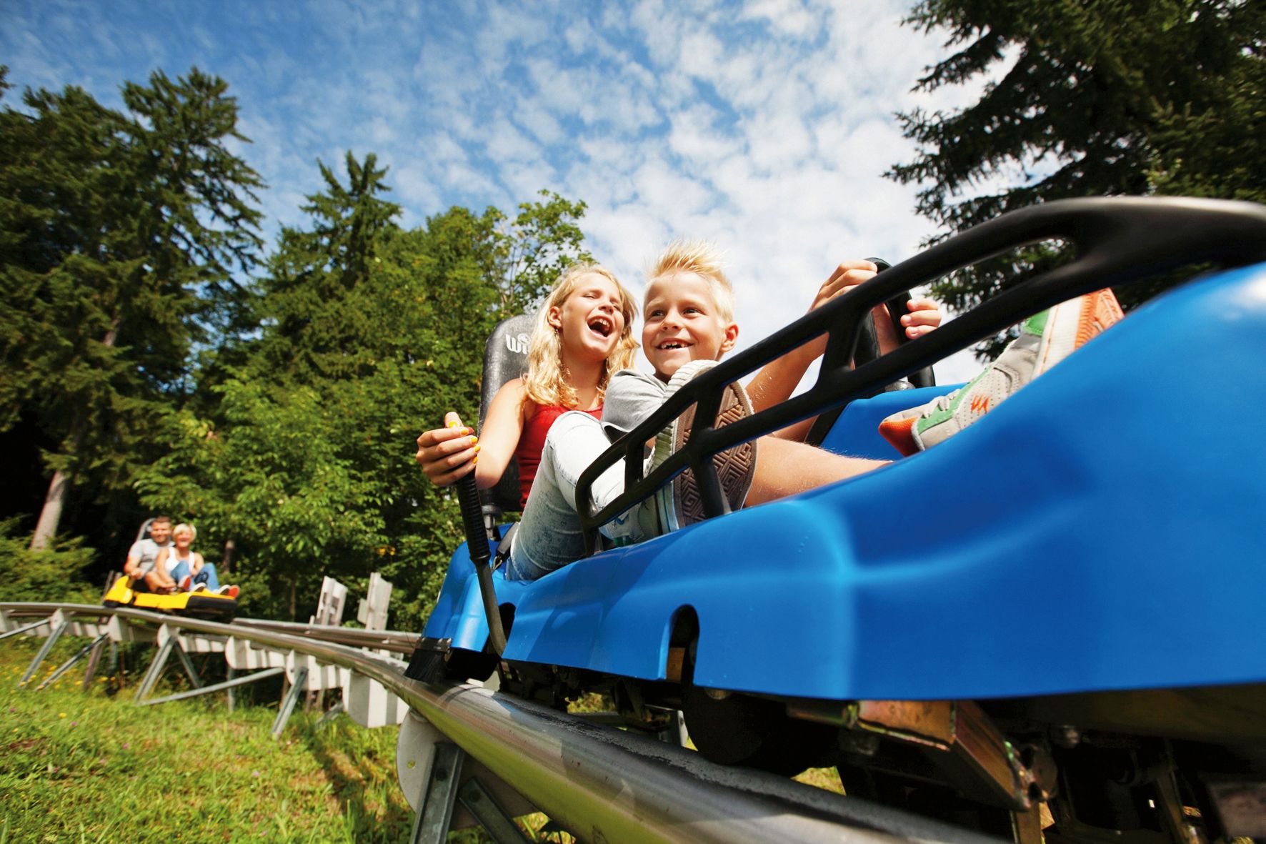 Zwei Kinder fahren lachend in einem blauen Sommerrodelbahn-Wagen durch einen Wald.