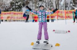 Ein fröhliches Kind genießt die ersten Schritte auf den Skiern und strahlt vor Freude. Umgeben von schneebedeckten Hängen und fröhlichen Skifahrern, vermittelt die Szene ein Gefühl von unbeschwerter Winterfreude.