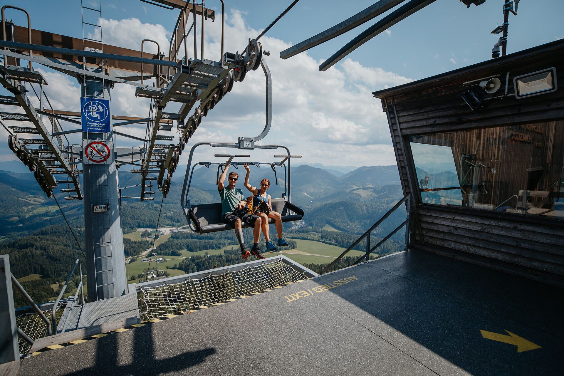 Ein Mann und eine Frau sind kurz davor, beim Sessellift der Annaberger Lifte am Annaberg auszusteigen. Im Hintergrund erstreckt sich eine Berglandschaft.