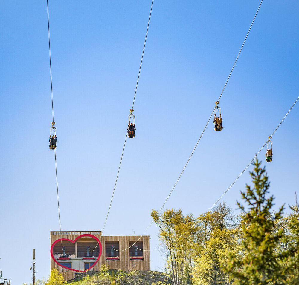 Mehrere Personen gleiten gleichzeitig auf der Zipline durch die Luft und bewegen sich vom Startbereich weg. Im Hintergrund ist ein Gebäude mit herzförmigem Fenster vor blauem Himmel zu sehen.