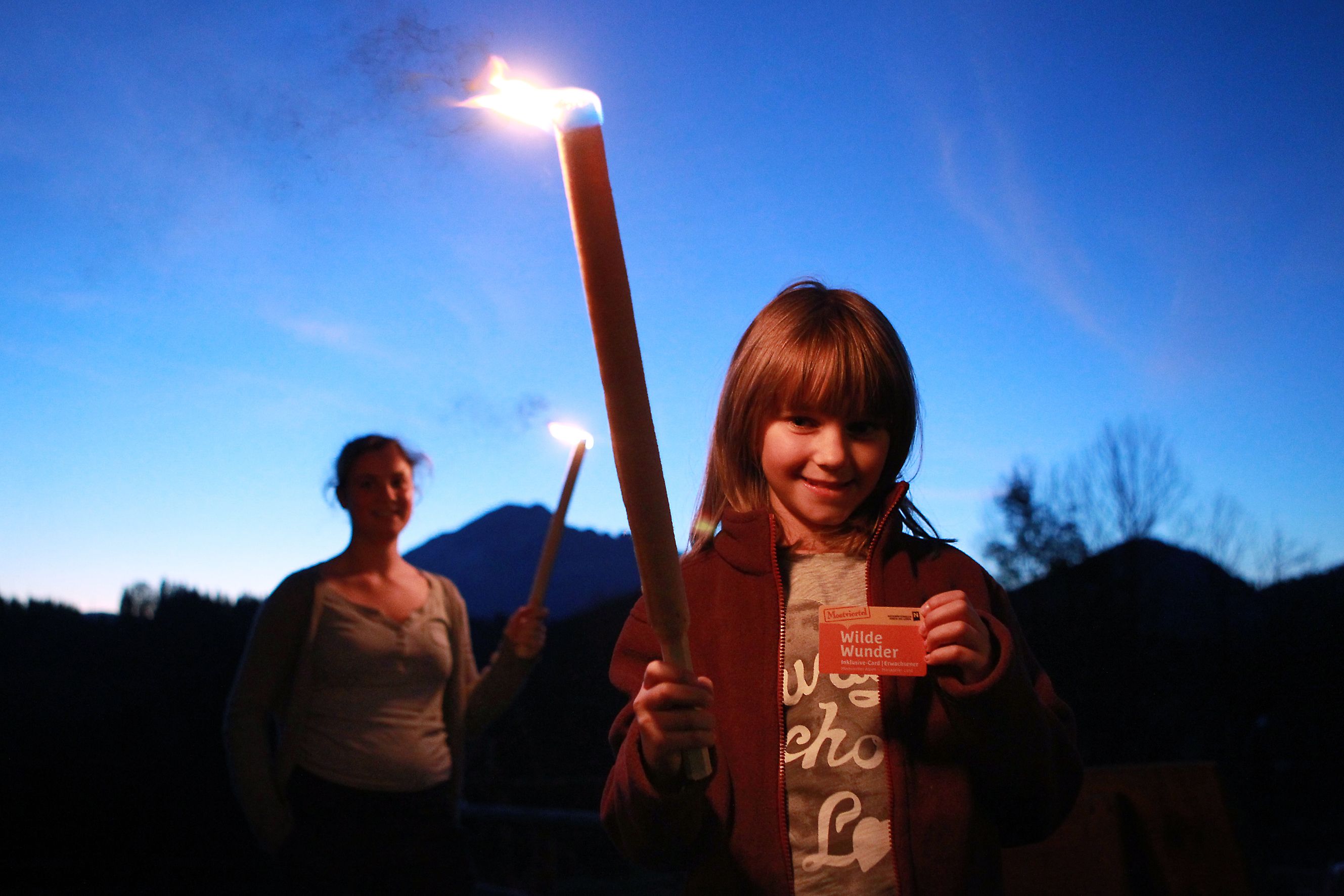A child holds a burning torch and a map, in the background a woman with a torch, against a mountain backdrop at dusk.