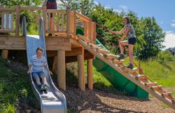 Eine Frau und zwei Kinder toben am Spielplatz. Der Junge rutscht über eine Rutsche und das Mädchen klettert über eine Holzleiter auf ein Podest.