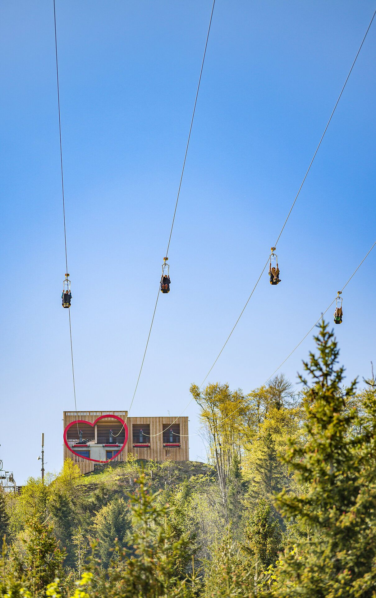 Mehrere Personen gleiten gleichzeitig auf der Zipline durch die Luft und bewegen sich vom Startbereich weg. Im Hintergrund ist ein Gebäude mit herzförmigem Fenster vor blauem Himmel zu sehen.