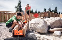 Ein Kind spielt zusammen mit seiner Mutter beim Wasserspielplatz. Während die Mutter das Wasser pumpt, spielt das Kind am anderen Ende der Wasserrinne.