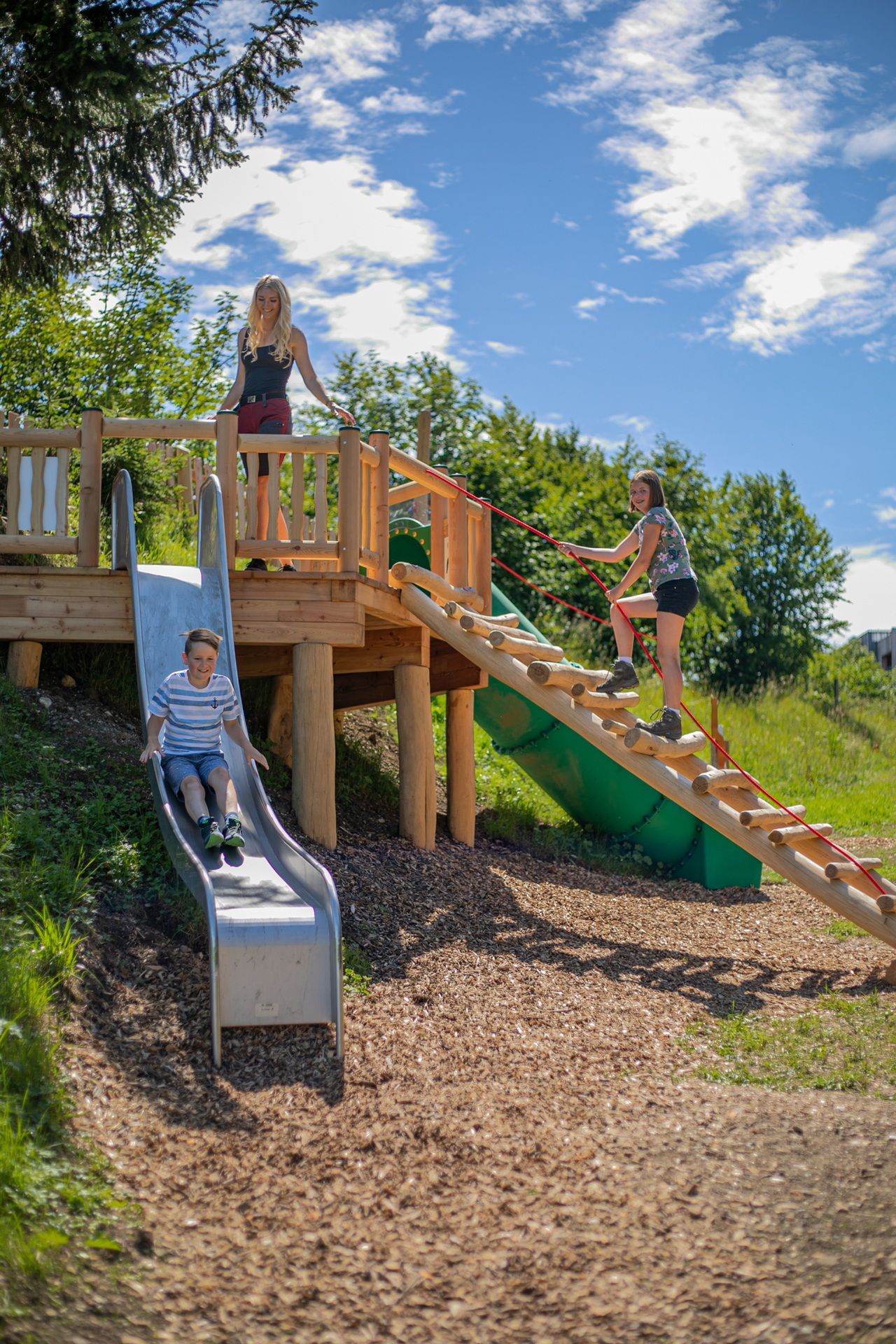 Eine Frau und zwei Kinder toben am Spielplatz. Der Junge rutscht über eine Rutsche und das Mädchen klettert über eine Holzleiter auf ein Podest.