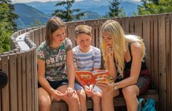 Eine Mutter mit zwei Kindern sitzt auf einer Bank und rätselt über etwas das im Entdeckerheft steht. Die Kinder rätseln ebenfalls mit.