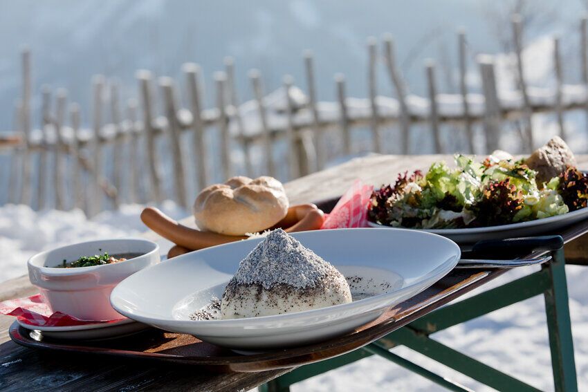 Inmitten der schneebedeckten Landschaft verströmt die rustikale Hütte ein einladendes Flair. Ein köstliches Gericht, umgeben von frischen Salaten und knusprigem Brot, verspricht Genuss und Gemütlichkeit. Hier wird der alpine Sommer in seiner schönsten Form erlebbar.