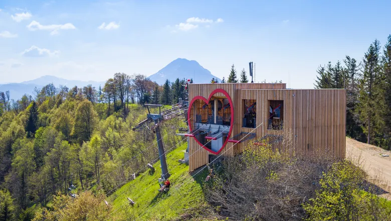 Holzgebäude mit Herzfenster und Seilbahn auf einem bewaldeten Hügel, Berg im Hintergrund.