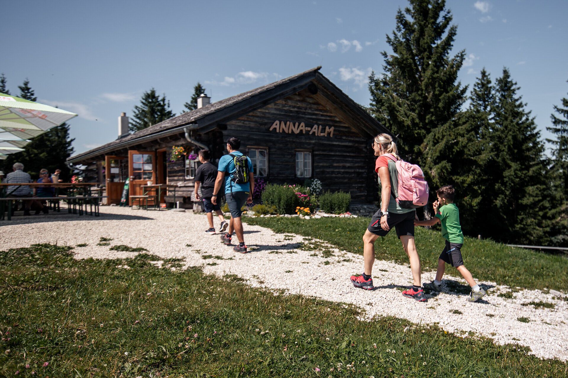 Eine Familie bestehend aus Mutter, Vater und zwei Söhnen ist auf dem Weg zur Anna-Alm am Annaberg. Im Hintergrund ist die Anna-Alm zu sehen, davor stehen mehrere Tische, an denen weitere Personen sitzen und die Atmosphäre genießen.