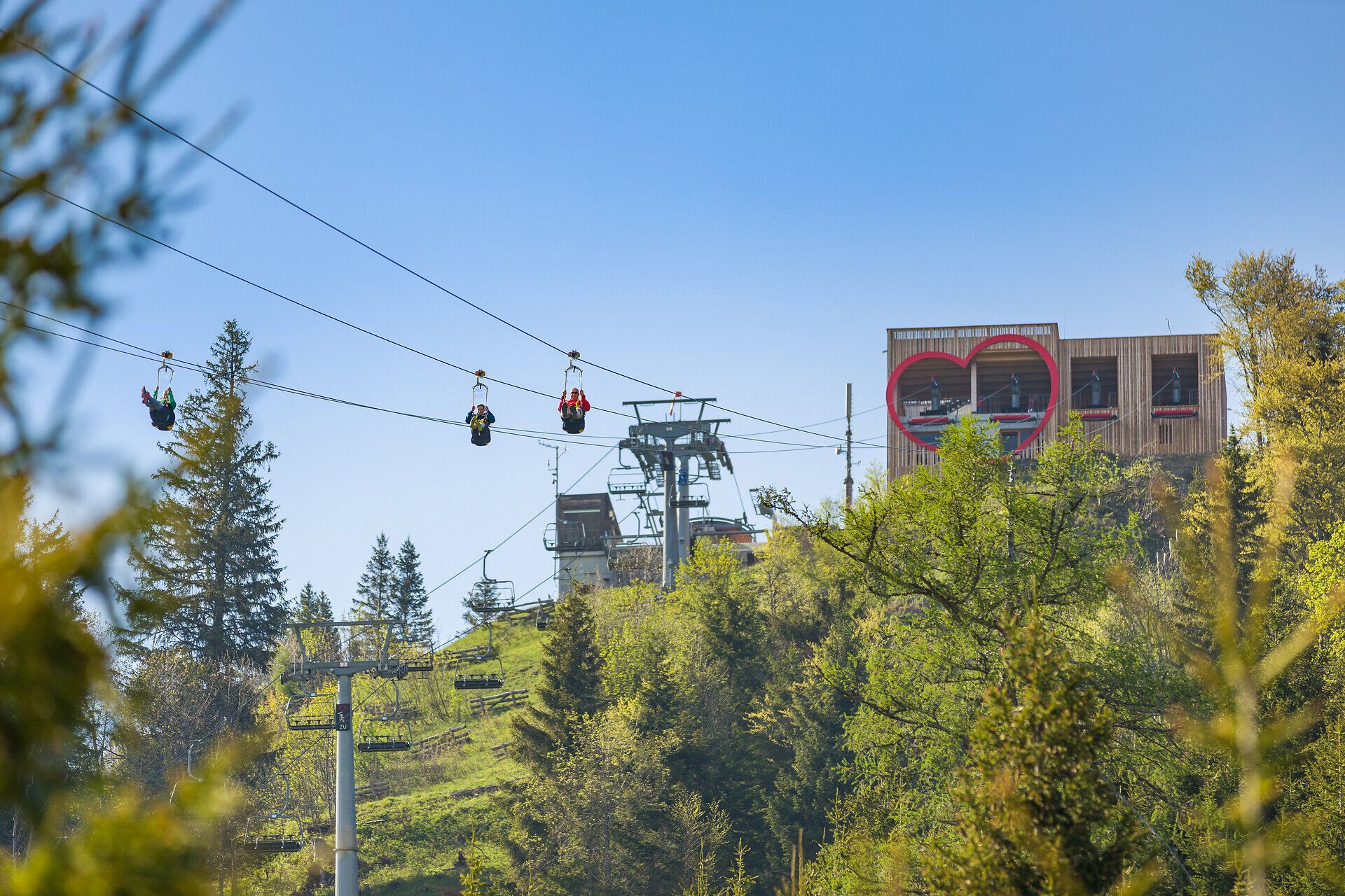 Mehrere Personen gleiten gleichzeitig auf der Zipline durch die Luft und bewegen sich vom Startbereich weg. Im Hintergrund ist ein Gebäude mit herzförmigem Fenster vor blauem Himmel zu sehen.