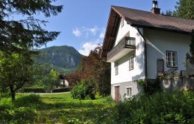 A white vacation home with balcony in front of a mountain landscape and green garden.