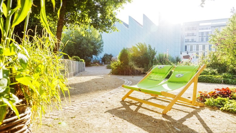 A sunny garden with green deckchairs and plants, with a modern building in the background.