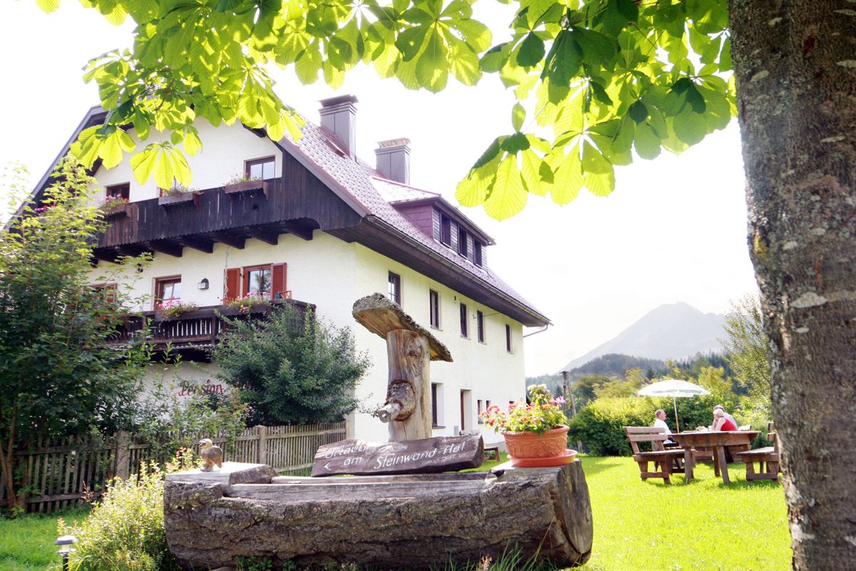 A traditional farmhouse with a wooden balcony, surrounded by green nature and a tree in the foreground.