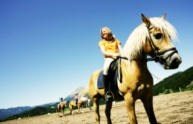 Eine Frau reitet auf einem braunen Pferd mit blonder M&auml;hne auf einem Reitplatz, umgeben von Bergen und blauem Himmel.