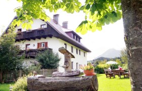 Ein traditionelles Bauernhaus mit Holzbalkon, umgeben von gr&uuml;ner Natur und einem Baum im Vordergrund.