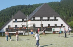 Kinder spielen Fußball auf einem Sportplatz vor einem Hotel in einer bewaldeten Gegend.