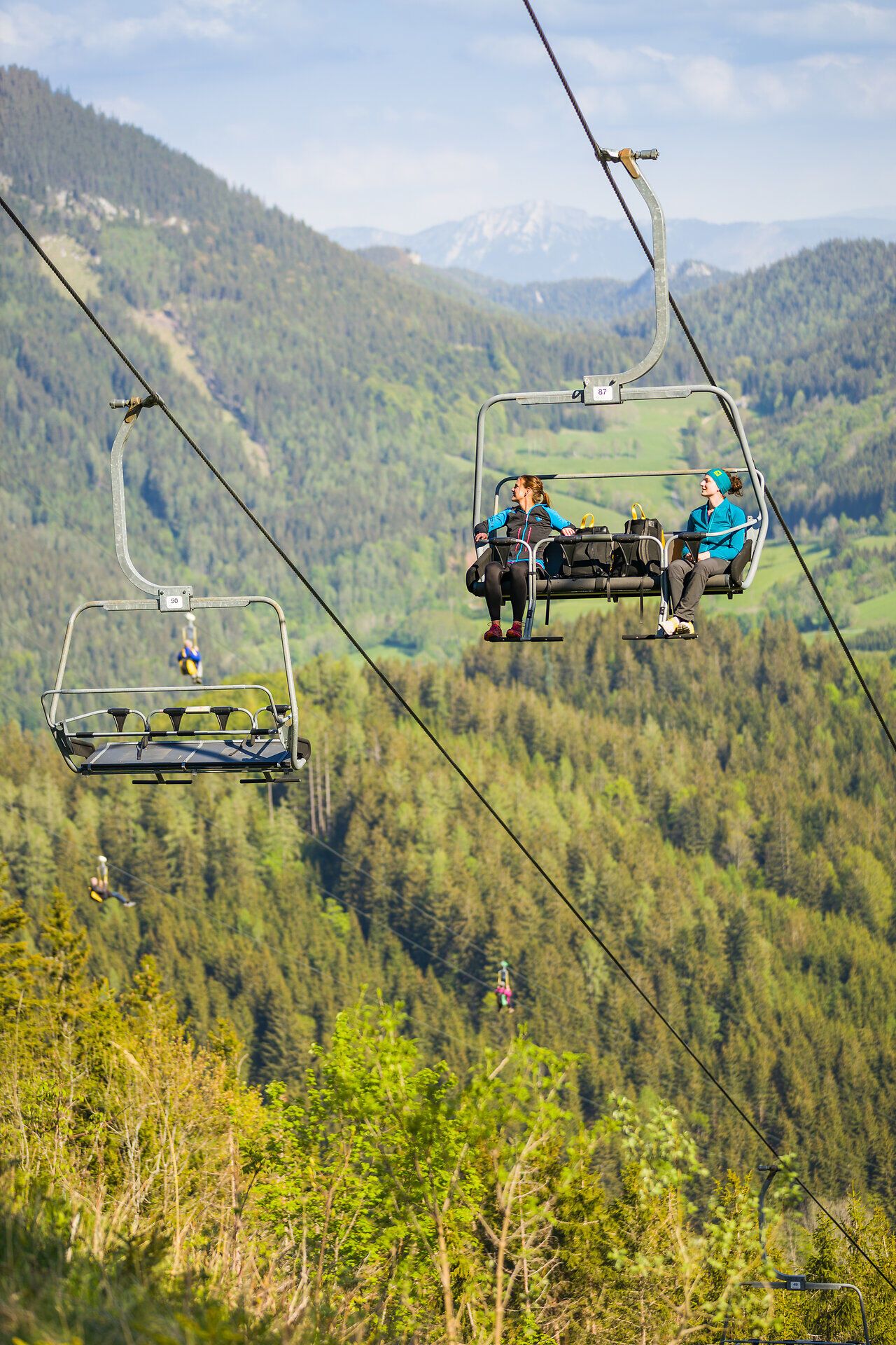 Zwei Personen sitzen auf einem Sessellift. Im Hintergrund ist die Zipline zu sehen, auf der mehrere Personen gleichzeitig durch die Luft gleiten. Außerdem sind Wälder und Berge der umliegenden Landschaft sichtbar.