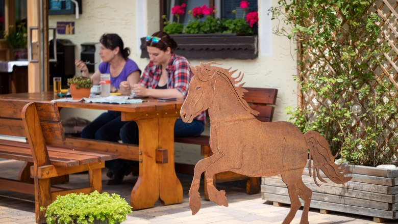 Two women sit at a wooden table outdoors and eat. In the foreground is a metal sculpture of a horse.