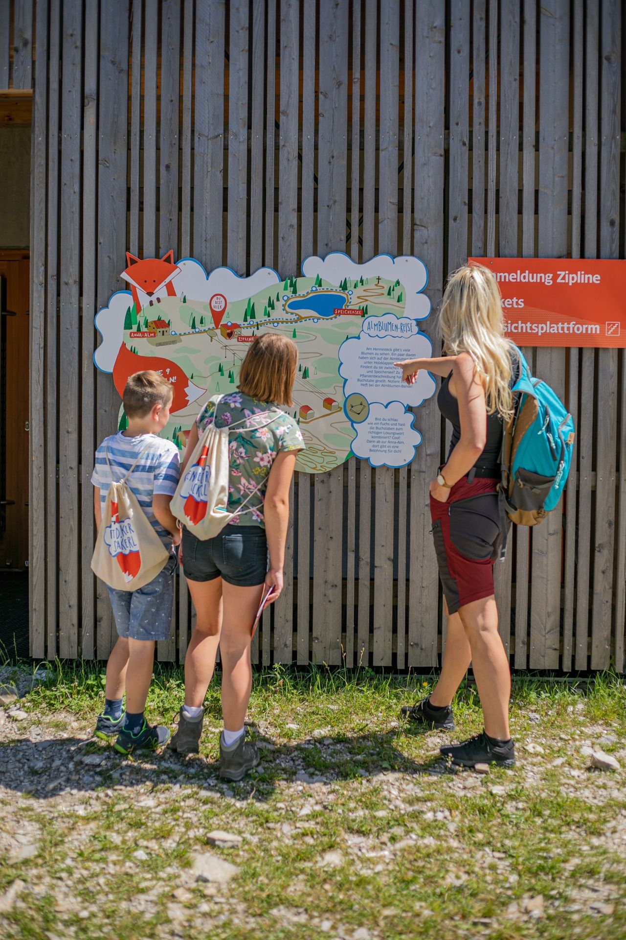 Eine junge Frau und zwei Kinder stehen vor dem Eingang zur Zipline und bewundern eine schöne große Informationstafel. Auf dieser ist Nikis Alm-Abenteuer zu sehen und eine große Informationssprechblase zu unseren Almblumen. Die junge Frau zeigt mit ihrem Finger auf die Tafel während die Kinder ganz interessiert zusehen.