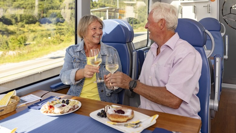 An elderly couple toast with wine glasses in one go, with food on the table.