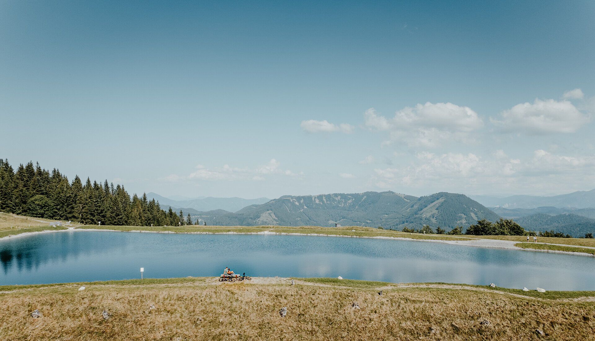 Ein Mann und eine Frau erkunden die Umgebung rund um den Speichersee am Hennesteck am Annaberg mit dem Fahrrad. Sie sind auf den Wegen rund um den See unterwegs und entdecken dabei die alpine Landschaft mit ihren Bergen, Wiesen und Aussichtspunkten.