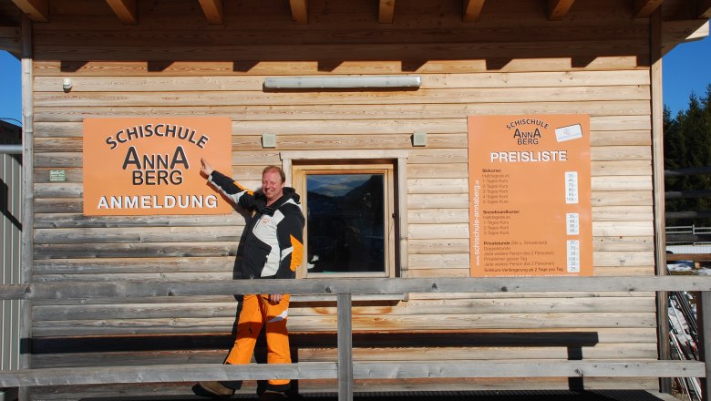 Ein Mann zeigt auf ein Schild der Schischule Annaberg vor einem Holzgebäude.