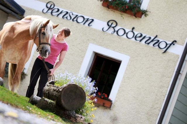 Eine Frau steht mit einem Pferd vor einem Gebäude mit der Aufschrift 'Pension Bodenhof'.