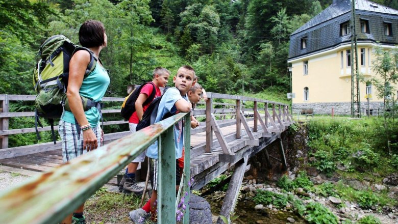 Eine Gruppe von Menschen steht auf einer Holzbrücke in einer grünen, bewaldeten Umgebung. Im Hintergrund ist ein gelbes Gebäude zu sehen.