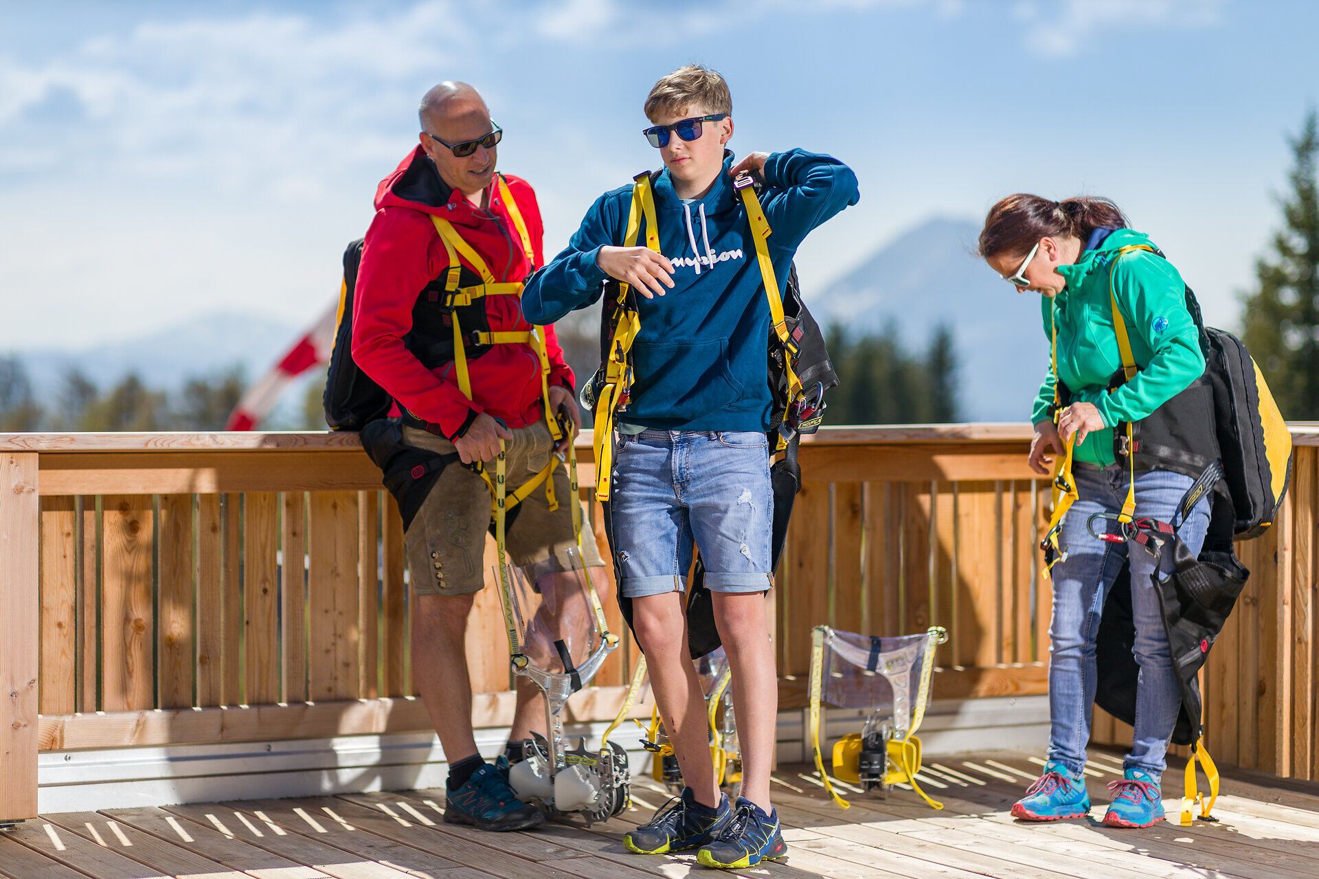 Drei Personen legen auf einer hölzernen Plattform die Sicherheitsgurte der Zipline an. Im Hintergrund sind Bäume und Berge der umliegenden Landschaft zu sehen.