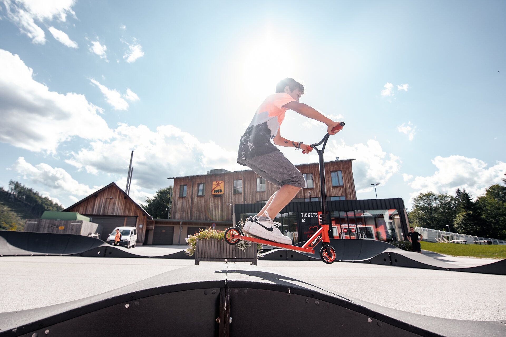 Ein Kind fährt mit einem Scooter auf dem Pumptrack in Annaberg an einem sonnigen Tag. Im Hintergrund ist das JUFA Hotel Annaberg zu sehen.