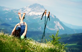 Person liegt auf einer Wiese mit Blick auf die Berge, Wanderstöcke daneben.