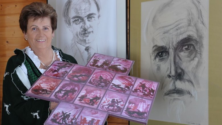 Woman in traditional costume holding artwork in front of two portraits.