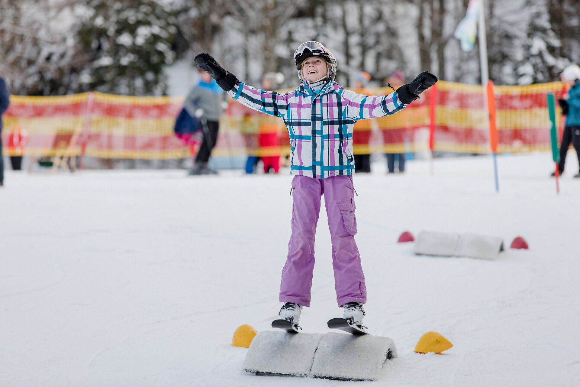 Ein fröhliches Kind genießt die ersten Schritte auf den Skiern und strahlt vor Freude. Umgeben von schneebedeckten Hängen und fröhlichen Skifahrern, vermittelt die Szene ein Gefühl von unbeschwerter Winterfreude.
